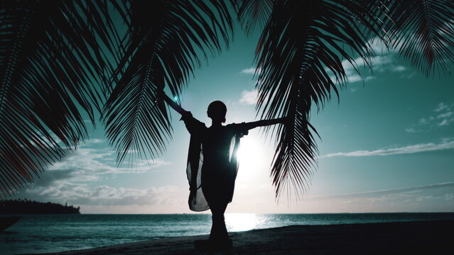Woman walking on night tropical beach. Silhouette of slim girl under the palm coconut tree raise hands up. Bright sky with sun rays reflected in water. Blue filter toning. Slow motion. Travel, tourism