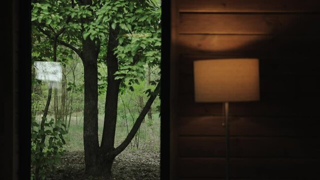 Window view from tiny wooden house, green forest outdoor, switched lamp with warm light near wooden wall, background.Closed window overlooking green garden, slow motion.