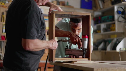 Senior and Junior Carpenters at Work, Young Apprentice and mentor Collaborating in Carpentry Workshop. person drilling wooden piece with drilling equipment