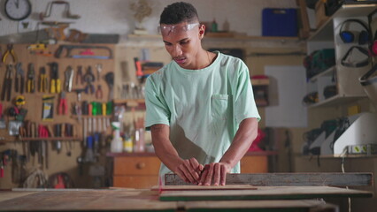 One Concentrated Young Black Brazilian Carpenter Engaged in Workshop Job, sawing piece of wood with industrial machine. Person engaged in job occupation