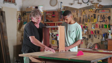 Senior carpenter guiding younger apprentice at carpentry workshop. Job occupation apprenticeship, mentor orienting and helping junior worker with craftmanship furniture