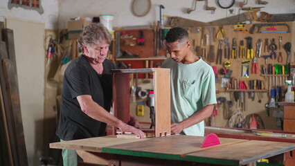 Senior carpenter guiding younger apprentice at carpentry workshop. Job occupation apprenticeship,...