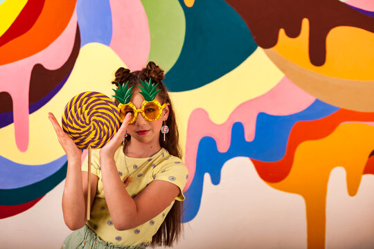 Happy child cover girl in pineapple glasses holds giant round lollipop at colorful wall, looking at camera. Perfect kid in yellow t-shirt with candy on stick. Summer sweet concept. Copy ad text space