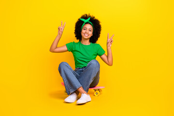 Full body photo of cute friendly funky girl sit skateboard demonstrate v-sign isolated on yellow color background