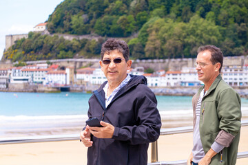 Two older men walking on the beach with a cell phone in their hand