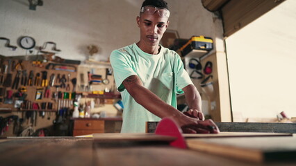 Young carpenter using saw machine to slice wood at workshop. Focused apprentice at carpentry artisan craftmanship store - obrazy, fototapety, plakaty