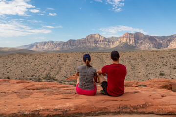 Couple sitting on edge of Calico Hills cliff with scenic view of limestone peaks Mount Wilson and Rainbow Mountain of Red Rock Canyon National Conservation Area, Mojave Desert, Las Vegas, Nevada, USA
