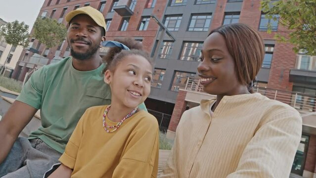 POV shot of cheerful African American parents and daughter in roller skates sitting outdoors on street, waving and smiling on camera during online video callPOV shot of cheerful African American paren