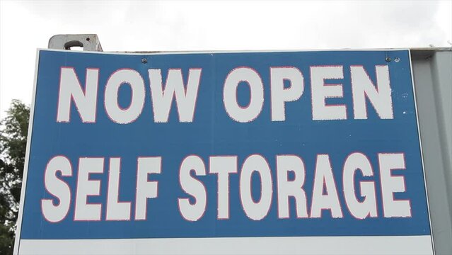 now open self storage sign in white writing caption text against blue background with sky and tree behind