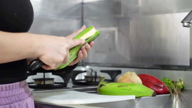 close-up of a woman's hands in the kitchen, peeling a green banana with a knife, preparing the food for cooking.