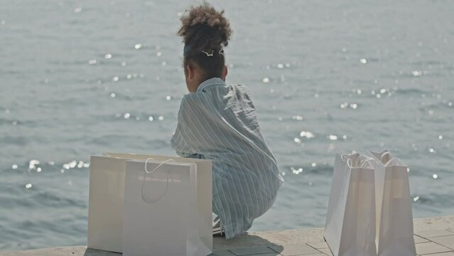 Back view of little African American girl sitting on riverside with shopping bags and looking at rippling waterBack view of little African American girl sitting on riverside with shopping bags and loo