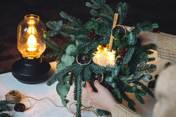 Young woman in knitted sweater doing handmade Christmas wreath from natural materials: fir tree branches, dry citrus slices, cones, cinnamon sticks. Wooden table, cozy light, home atmosphere