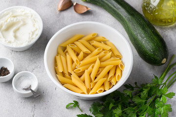Penne pasta, zucchini, cream cheese, garlic, parsley, olive oil, salt, pepper for delicious vegetarian pasta on a gray textured background, top view
