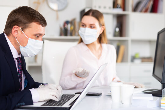 Portrait of busy entrepreneur in medical face mask and latex gloves working with female coworker in office. Concept of precautions and social distancing in coronavirus pandemic..