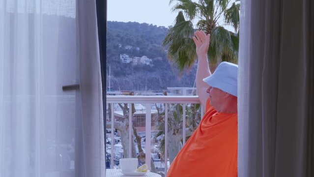 mature man in orange t-shirt, senior of 60 years old sitting on balcony, enjoys view outskirts palm trees, yachts, resort town port Soller on island Mallorca, enjoying journey, vacation, resort place