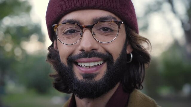 Close up of smiling young Caucasian millennial in winter clothes looking at camera positive attitude. Happy alternative man with hipster beard and earring posing outdoors. Blurred park background.