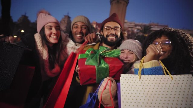 Evening winter smiling multiracial group friends showing bags gifts to camera. Happy young millennial pose for video outdoor city at night. People enjoying christmas vacations and black Friday sales.