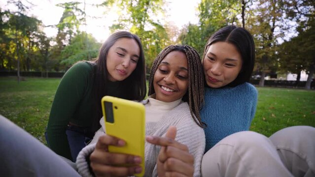 Three young multiracial women using smiling cell phone in park. Three teenage girls amusingly gossiping on a smartphone app. Millennial people connected through online platforms and social networks. 