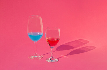 Minimal scene wine glass with red wine and blue cocktail on sunny day. Summer concept drinks and shadows on pink background.