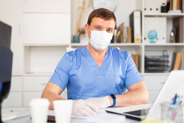 Fototapeta na wymiar Portrait of male doctor in face mask and gloves working on laptop consulting patient online, telemedicine concept