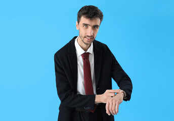 Young man in suit looking at wristwatch on blue background