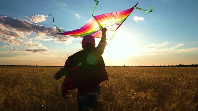 Active Child runs with kite in sun. Happy girl runs in field of wheat, plays with toy kite. Kite hovers flies in hand of kid. Girl with toy kite. Daughter plays at sunset, dreams of flying, traveling
