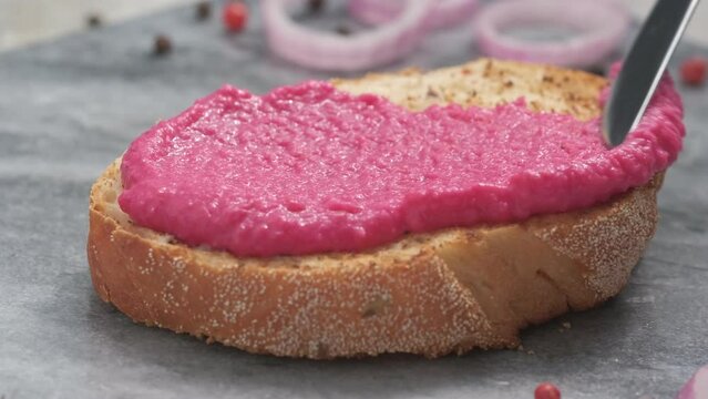 Spreading horseradish and beetroot paste spread with knife on top of toasted bread slice close up