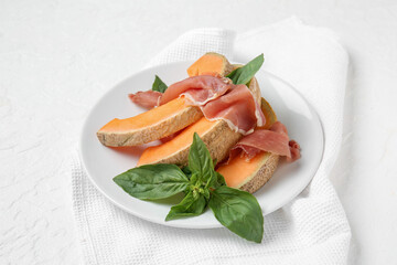 Plate with tasty melon, prosciutto and basil on light background