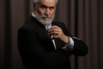 Senior man in formal suit holding glass of whiskey with ice cubes on brown background