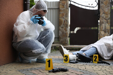 Naklejka na ściany i meble Criminologist taking photo of evidence at crime scene with dead body near house