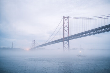 Fototapeta na wymiar View of 25 de Abril Bridge famous tourist landmark of Lisbon connecting Lisboa and Almada in heavy fog mist wtih yacht boats passing under. Lisbon, Portugal