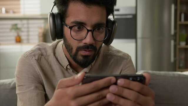 Shocked amazed Arabian man Indian guy watch video stream online sport match social media addict focused male in headphones looking at mobile phone screen watching movie on smartphone at home kitchen
