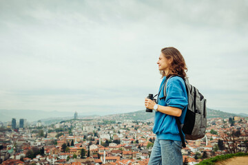 A young adult smiling female traveler looks through binoculars at a European city with orange roofs...