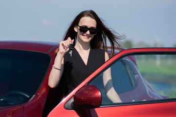 Beautiful young girl in a black overalls and sunglasses stands near the open driver's door of a red car holding on to it and holding the car keys in her hand