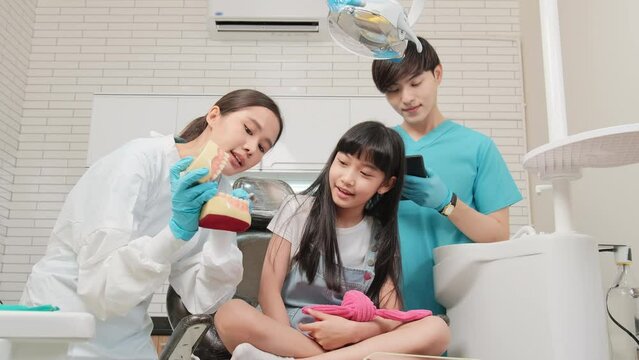 Asian female pediatric dentist and assistant demonstrate toothbrush to little girl with teeth model in dental clinic, well-being hygiene, and professional orthodontic healthcare work in kid hospital.