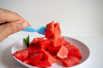 Fresh watermelon ready to eat using a blue fork on a white plate with a white background
