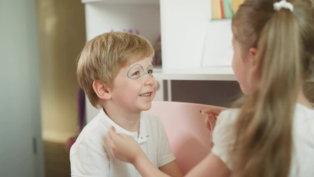 Girl paints glasses on younger boy face and children smile