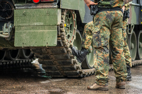 Two soldiers in camouflage uniform standing during a break under the rain near the tracks of an armored dirty tank, only the trousers and the boots are visible 
