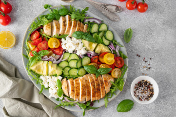 Salad with roasted chicken, avocado, cherry tomatoes, cucumbers, arugula and feta cheese on a plate on a gray concrete background. Healthy food.