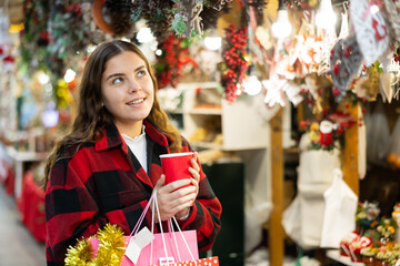 Portrait of happy attractive young girl in light plaid jacket visiting traditional Christmas fair in European city, walking among shopping stalls with red paper cup of hot coffee