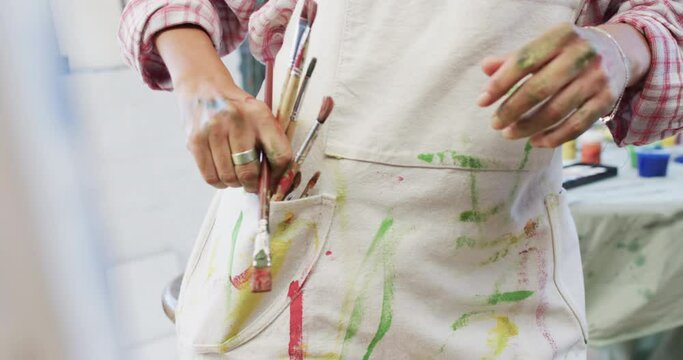 Midsection of biracial female artist putting paint brushes in pocket in studio, slow motion