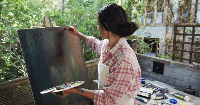 Biracial female artist in apron painting on canvas in sunny garden, slow motion