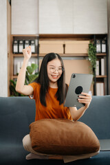 Obraz premium Attractive happy young Asian student studying at the college library, sitting at the sofa, using a laptop computer, tablet and headphones having a video chat.