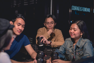 A handsome guy in brown long sleeves is staring blankly ahead, holding his coffee, leaning on the back chair while his other friends are talking. Window glass, neon sign and a dark background is seen