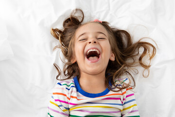 Little girl lying on white bed laughing with open mouth and closed eyes