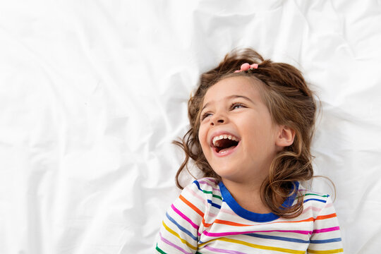 Portrait of laughing little girl lying on white bed looking up