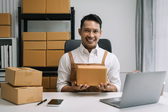 Young Asian man holding parcels and looking to camera, standing among several boxes in home office.