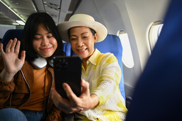 Smiling middle aged woman and daughter sitting in passenger airplane and taking picture, waiting...