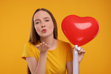 Young woman holding red heart shaped balloon and blowing kiss on yellow background