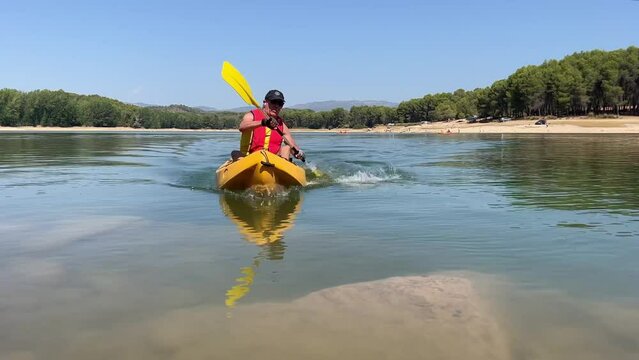 Middle-aged white man in a yellow kayak rowing in a lake. He is wearing a red jacket and a black cap. Sunny day. Summer. Jerica, Castellon. Spain.